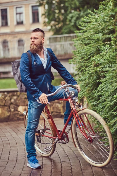 Бородатый мужчина со стильной стрижкой, одетый в повседневную одежду с рюкзаком, сидящий на ретро-велосипеде в городском парке . — стоковое фото