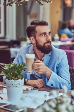 Şık bir saç kesimi ile şık bir sakallı erkek portresi bir kafe mekanlarda oturan bir kapuçino, bir bardak içer.