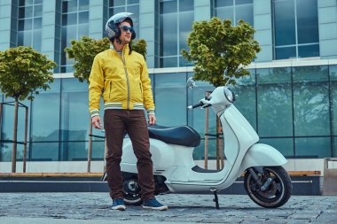 Yakışıklı erkek kahverengi pantolon ve güneş gözlüğü ve kask, bir gökdelen karşı klasik İtalyan scooter yakın duran sarı ceket giyiyor.