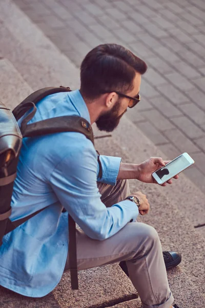 Tourist with a full beard and haircut, wearing casual clothes with a backpack, texting on a smartphone, sitting on a step in an antique street. — Stock Photo, Image