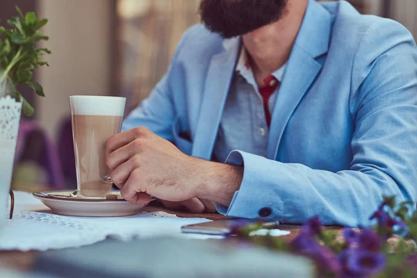 Retrato de um homem barbudo na moda com um corte de cabelo elegante, segura um copo de um cappuccino, sentado em um café ao ar livre . — Fotografia de Stock
