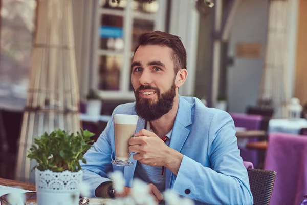 Retrato de um homem barbudo na moda com um corte de cabelo elegante, bebe um copo de um cappuccino, sentado em um café ao ar livre . — Fotografia de Stock