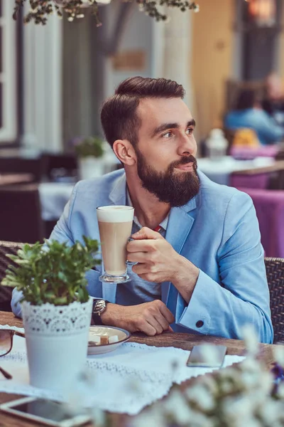 Портрет модного бородатого чоловіка зі стильною стрижкою, п'є келих капучино, сидячи в кафе на відкритому повітрі . — стокове фото