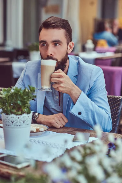 Πορτρέτο ενός μόδας γενειοφόρου άνδρα με ένα μοντέρνο κούρεμα, πίνει ένα ποτήρι καπουτσίνο, κάθεται σε μια υπαίθρια καφετέρια. — Φωτογραφία Αρχείου