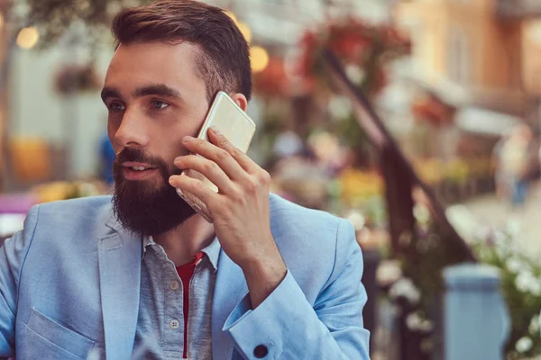 Retrato de cerca de un hombre de negocios barbudo de moda con un corte de pelo elegante, hablando por teléfono, bebe un vaso de jugo, sentado en un café al aire libre . —  Fotos de Stock