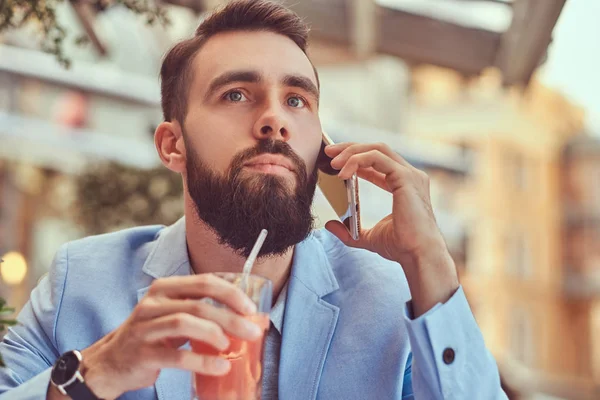 Retrato de cerca de un hombre de negocios barbudo de moda con un corte de pelo elegante, hablando por teléfono, bebe un vaso de jugo, sentado en un café al aire libre . —  Fotos de Stock