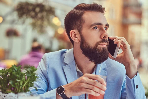 Retrato de cerca de un hombre de negocios barbudo de moda con un corte de pelo elegante, hablando por teléfono, bebe un vaso de jugo, sentado en un café al aire libre . —  Fotos de Stock