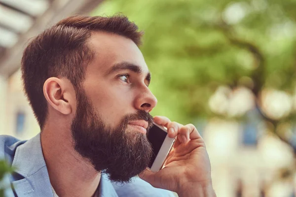Крупним планом портрет модного бородатого чоловіка зі стильною стрижкою, розмовляючи по телефону, сидячи в кафе на відкритому повітрі . — стокове фото