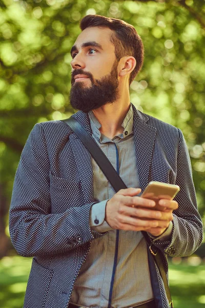 Portret van een bebaarde man met een stijlvol kapsel in casual kleding, schrijft een bericht op de telefoon, staat in een park. — Stockfoto