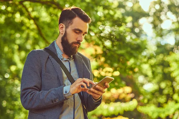 Портрет бородатого мужчины со стильной стрижкой в повседневной одежде, пишет сообщение по телефону, стоит в парке . — стоковое фото