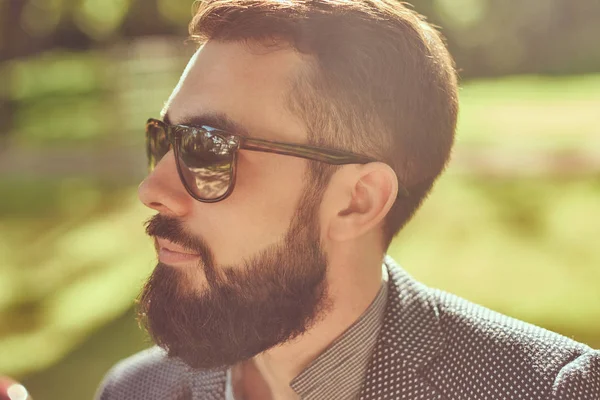 Close-up portrait of a bearded male with a stylish haircut, in a jacket and sunglasses, sitting on a bench in a park. — Stock Photo, Image