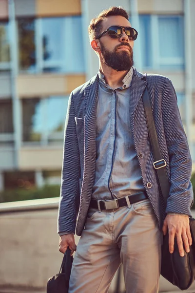 Retrato de um trabalhador barbudo vestindo roupas casuais e óculos de sol, com uma bolsa, em pé em uma rua da cidade contra um arranha-céu . — Fotografia de Stock