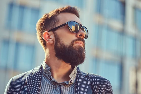 Portrait en gros plan d'un homme barbu portant des vêtements décontractés et des lunettes de soleil, debout dans une ville contre un gratte-ciel . — Photo