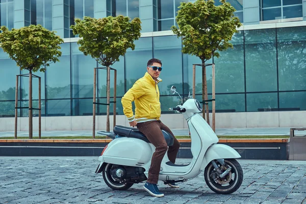 Hombre guapo con pantalones marrones chaqueta amarilla y gafas de sol, sentado en un clásico scooter italiano blanco contra un rascacielos . —  Fotos de Stock