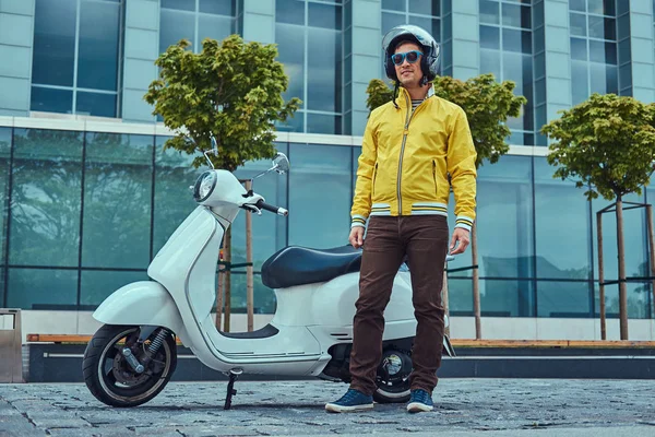 Hombre guapo con pantalones marrones y chaqueta amarilla, en gafas de sol y casco, de pie cerca de la clásica scooter italiana contra un rascacielos . — Foto de Stock