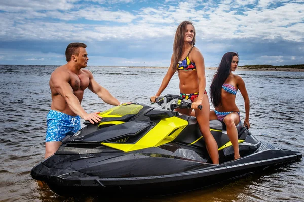 Two sexy girls in swimsuit and shirtless muscular male, have fun with a jet ski on a sea. Stock Picture
