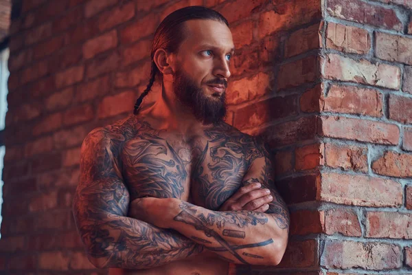 Un guapo hombre sin camisa tatuado con un corte de pelo elegante y barba, de pie con los brazos cruzados, apoyado contra una pared de ladrillo en una habitación con un interior loft . —  Fotos de Stock