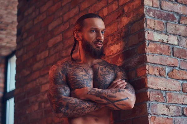 Un guapo hombre sin camisa tatuado con un corte de pelo elegante y barba, de pie con los brazos cruzados, apoyado contra una pared de ladrillo en una habitación con un interior loft . — Foto de Stock