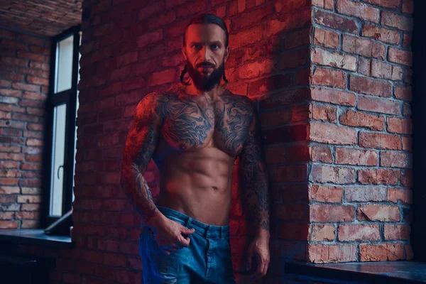 Um belo homem sem camisa tatuado com um corte de cabelo elegante e barba, de pé contra uma parede de tijolo em uma sala com um interior loft . — Fotografia de Stock