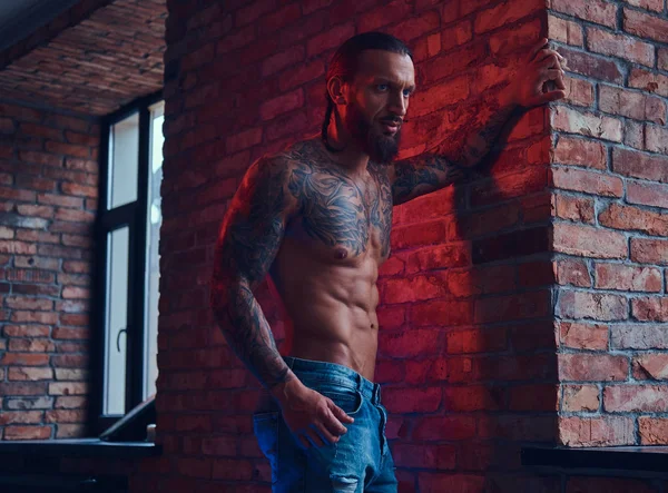 Um belo homem sem camisa tatuado com um corte de cabelo elegante e barba, de pé contra uma parede de tijolo em uma sala com um interior loft . — Fotografia de Stock