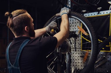 Bir bisiklet tekerleği bir tamir dükkanında çalışan yakışıklı Kızıl saçlı erkek kot pantolon coverall içinde. Bir işçi Bisiklet lastik bir atölyede kaldırır..