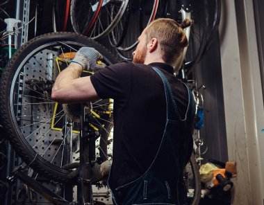 Bir bisiklet tekerleği bir tamir dükkanında çalışan yakışıklı Kızıl saçlı erkek kot pantolon coverall içinde. Bir işçi Bisiklet lastik bir atölyede kaldırır..