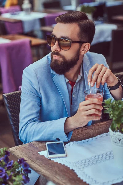 Portret van een modieuze bebaarde man met een stijlvol kapsel, houdt een glas voor een cappuccino, zittend in een café in de buitenlucht. — Stockfoto