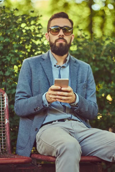 Retrato de um macho barbudo com um corte de cabelo elegante em roupas casuais, escreve uma mensagem ao telefone enquanto se senta em um banco em um parque . — Fotografia de Stock