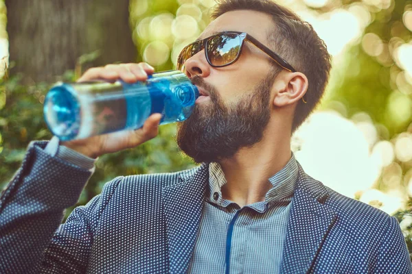 Vousatý muž muž pití studené vody venku, sedí na lavičce v městském parku. — Stock fotografie