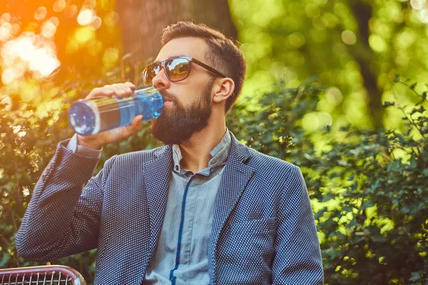 Vousatý muž muž pití studené vody venku, sedí na lavičce v městském parku. — Stock fotografie