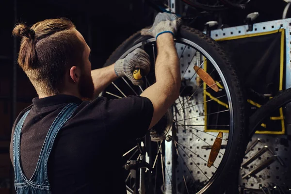 Красивый рыжий мужчина в джинсовом комбинезоне, работает с велосипедным колесом в ремонтной мастерской. Рабочий снимает велосипедную шину в мастерской . — стоковое фото