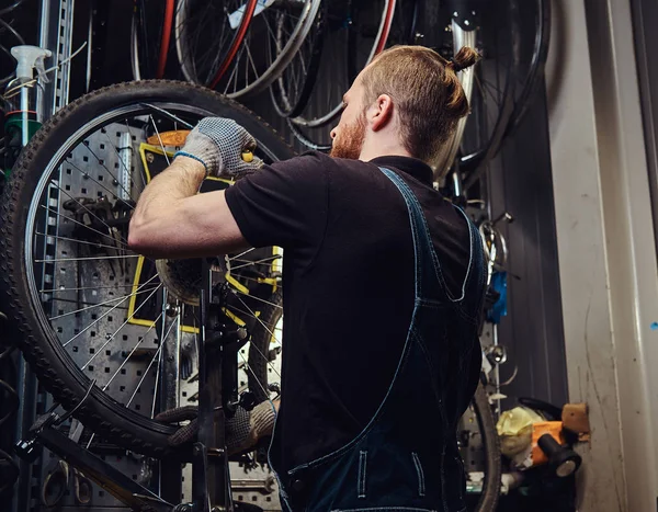 Homem ruivo bonito em um macacão jeans, trabalhando com uma roda de bicicleta em uma oficina de reparação. Um trabalhador remove o pneu da bicicleta em uma oficina . — Fotografia de Stock
