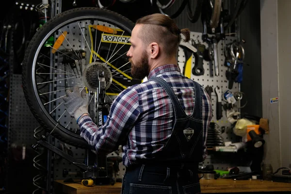 Красивый стильный мужчина в фланелевой рубашке и джинсовом комбинезоне, работает с велосипедным колесом в ремонтной мастерской . — стоковое фото