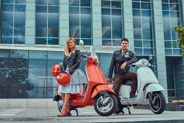 Aantrekkelijke romantisch paar, een knappe man en sexy vrouw, permanent met twee retro Italiaanse scooters tegen een wolkenkrabber. — Stockfoto