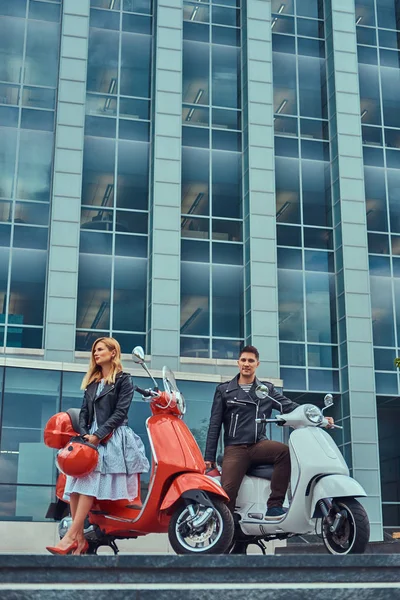 Casal romântico atraente, um homem bonito e sexy feminino, de pé com duas scooters retro italiano contra um arranha-céu . — Fotografia de Stock