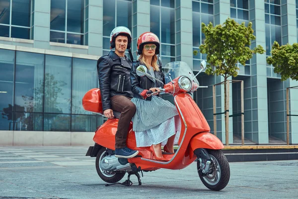 Um casal atraente, um homem bonito e sexy feminino montando juntos em uma scooter retro vermelho em uma cidade . — Fotografia de Stock