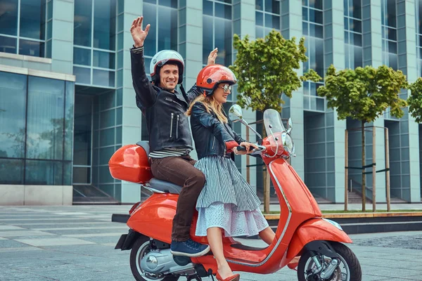 Atractiva pareja feliz, un hombre guapo y sexy hembra cabalgando juntos en un scooter retro rojo en una ciudad . — Foto de Stock