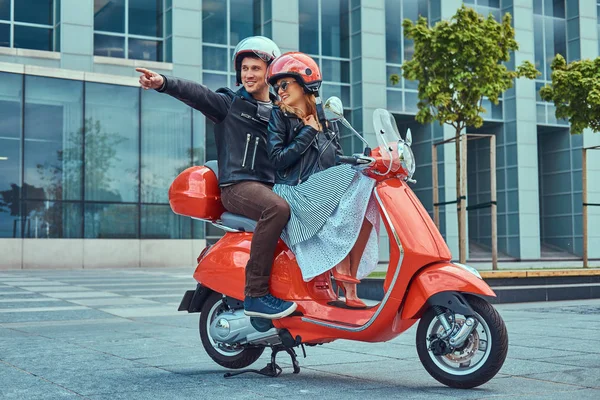 Atractiva pareja feliz, un hombre guapo y sexy hembra cabalgando juntos en un scooter retro rojo en una ciudad . — Foto de Stock