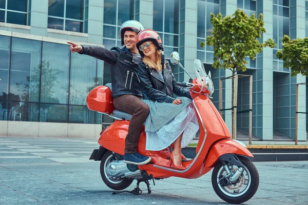 Attraktives glückliches Paar, ein gutaussehender Mann und eine sexy Frau, die zusammen auf einem roten Retro-Roller in einer Stadt fahren. — Stockfoto