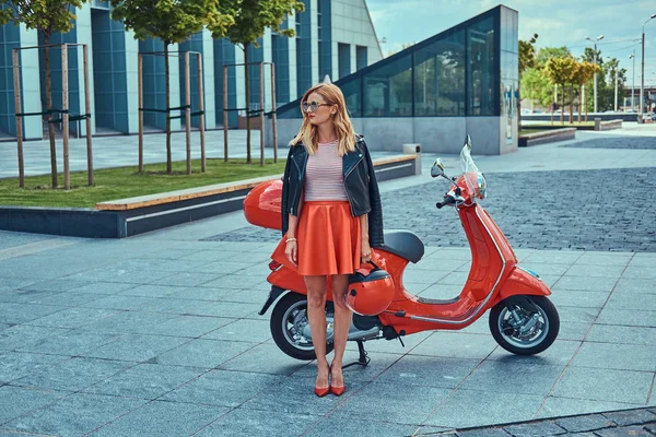 Sexy stylische blonde Mädchen trägt einen roten Rock und schwarze Lederjacke und Sonnenbrille, hält einen Helm und steht in der Nähe klassischer italienischer Roller gegen einen Wolkenkratzer. — Stockfoto