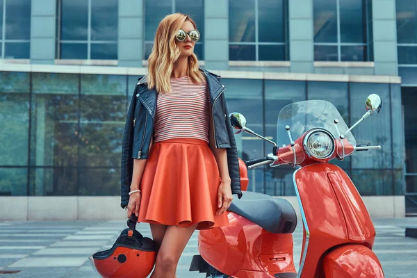 Sexy chica rubia elegante con una falda roja y chaqueta de cuero negro y gafas de sol, sosteniendo un casco y de pie cerca de la clásica scooter italiana contra un rascacielos . —  Fotos de Stock