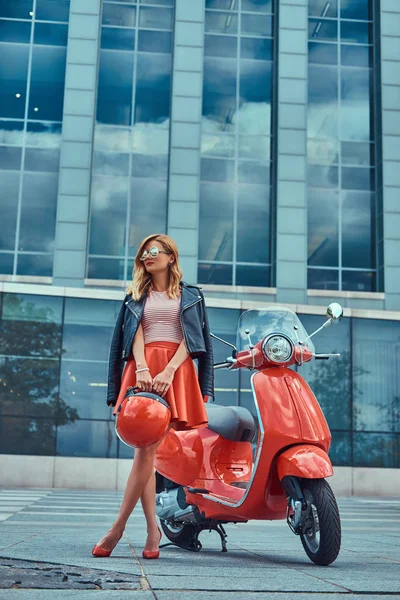 Sexy chica rubia elegante con una falda roja y chaqueta de cuero negro y gafas de sol, sosteniendo un casco y de pie cerca de la clásica scooter italiana contra un rascacielos . —  Fotos de Stock