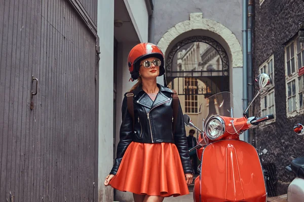 Menina loira sexy vestindo roupas elegantes em óculos de sol e capacete, de pé em uma rua estreita velha com duas scooters retro . — Fotografia de Stock