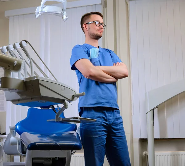 Bonito dentista masculino barbudo em uniforme azul e óculos, de pé com os braços cruzados em uma clínica odontológica . — Fotografia de Stock