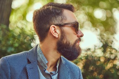 Yakın çekim bir ceket ve güneş gözlüğü bir şehir parkta bir bankta oturmuş, şık bir saç kesimi ile sakallı bir erkek portresi.