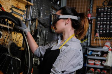 İş giyisi, önlük ve gözlük, güzel kadın bir bisiklet tekerleği bir atölyede onarır.