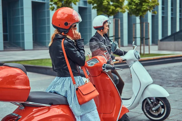 An attractive romantic couple, a handsome man and sexy female sitting on retro Italian scooters against a skyscraper. A man waiting while girl talking by phone.