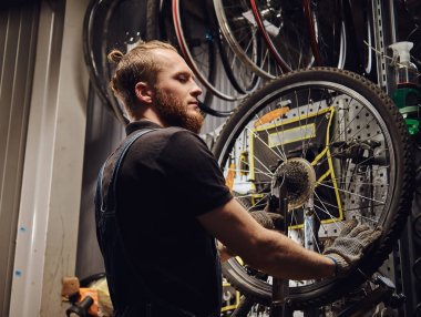 Bir bisiklet tekerleği bir tamir dükkanında çalışan bir yakışıklı Kızıl saçlı erkek kot pantolon coverall içinde. Bir işçi Bisiklet lastik bir atölyede kaldırır..
