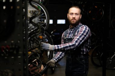 Bir bisiklet tekerleği bir tamir dükkanında çalışan bir fanila coverall, tişört ve kot giyen bir yakışıklı şık erkek. Bir işçi bir anahtarı kullanarak bir atölyede bisikletle tekerlek bağlar.