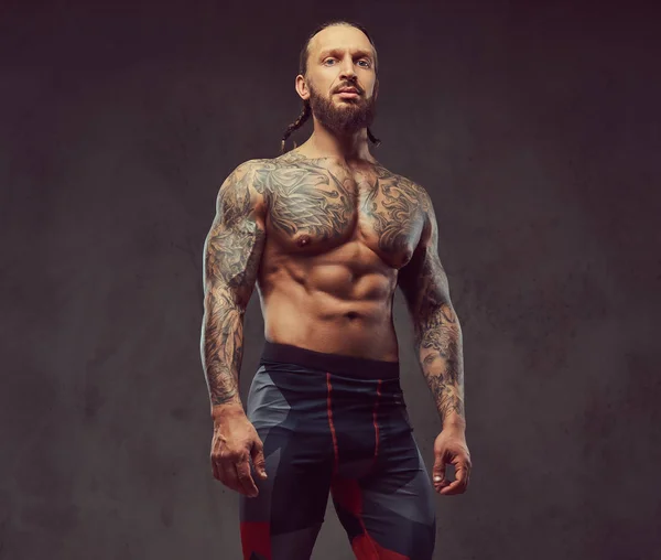 スポーツ ズボンでスタイリッシュな散髪と筋肉裸髭刺青男性の肖像画 暗い背景に分離 — ストック写真